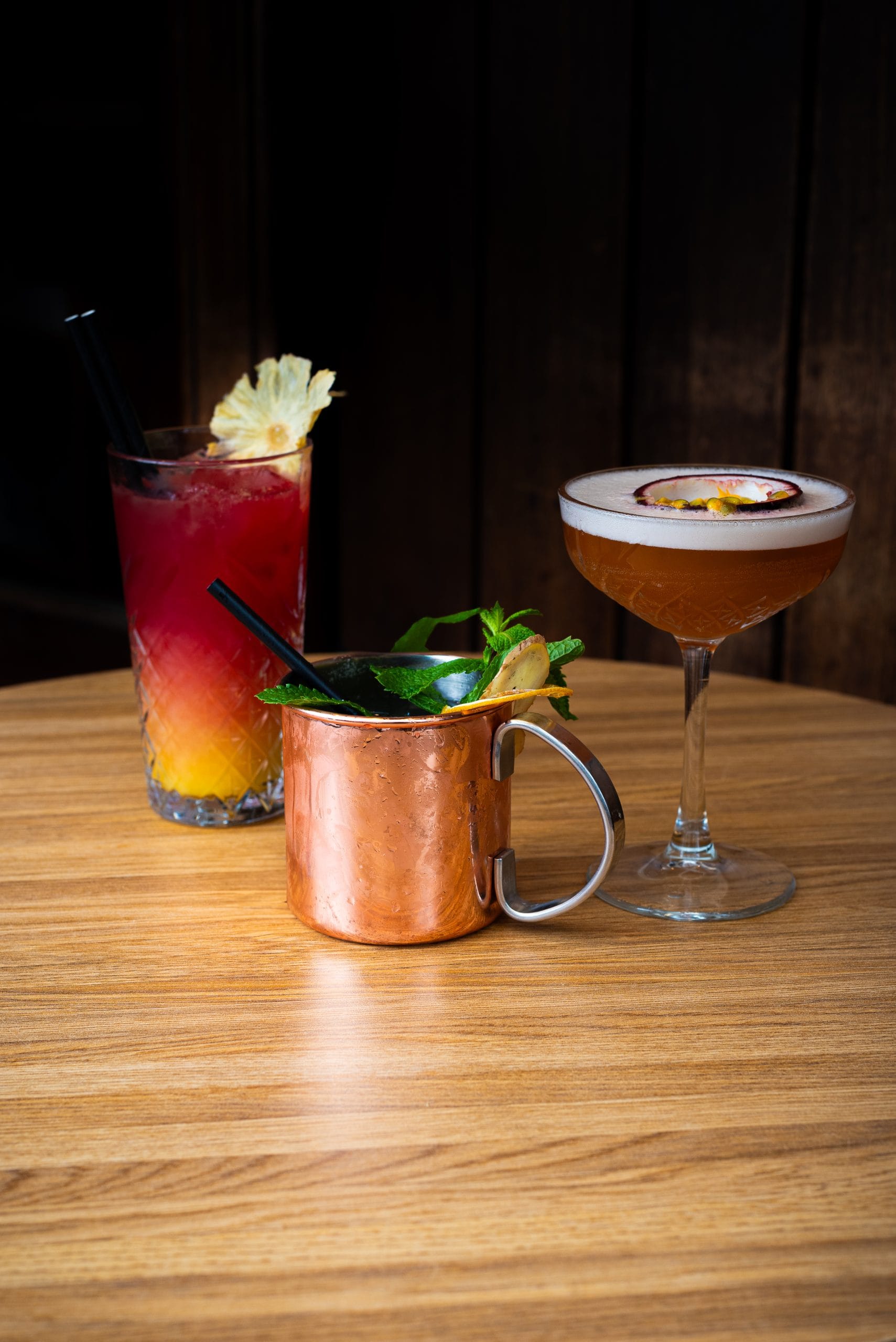 Drie cocktails op een houten tafel: een longdrink, een mule in koperen beker en een Pornstar Martini